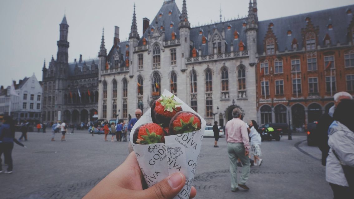 Una mano tiene un cono di carta di fragole ricoperte di cioccolato in una vivace piazza storica europea.
