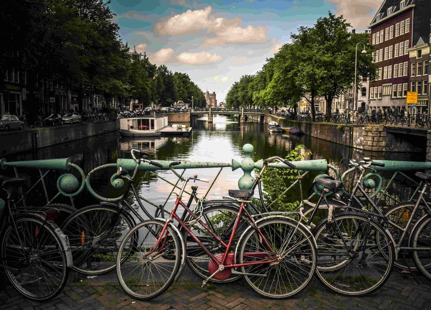 Biciclette parcheggiate contro una ringhiera verde su un ponte che si affaccia su un canale cittadino fiancheggiato da alberi ed edifici.