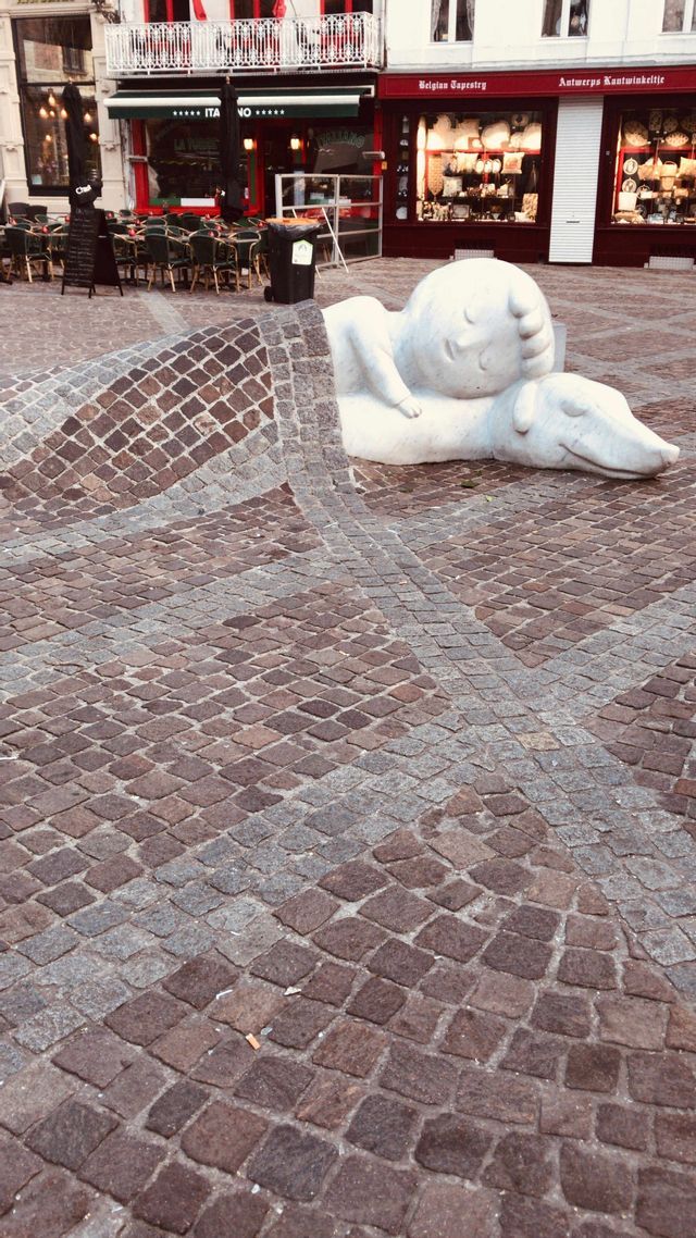 Una scultura bianca di un bambino che dorme su un cane, con un selciato di ciottoli che funge da coperta su di loro in una piazza della città.