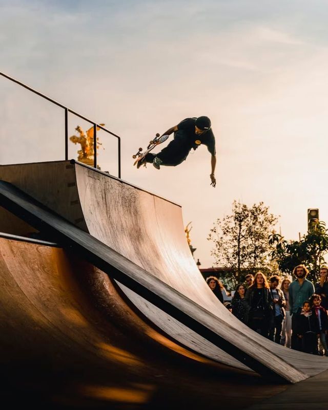 Uno skateboarder in tenuta nera è in aria sopra una rampa half-pipe in legno in uno skatepark al tramonto.