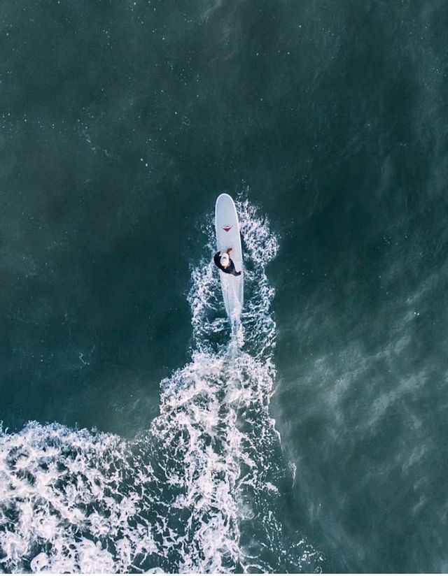 Veduta aerea dall'alto di un surfista su una tavola bianca che cavalca un'onda in acque scure, lasciando una scia bianca.