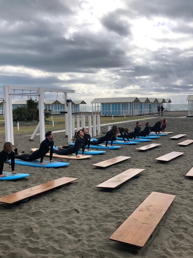Un gruppo WeRoad in muta pratica una posa di riscaldamento su tavole da surf su una spiaggia sabbiosa, sotto un cielo coperto.