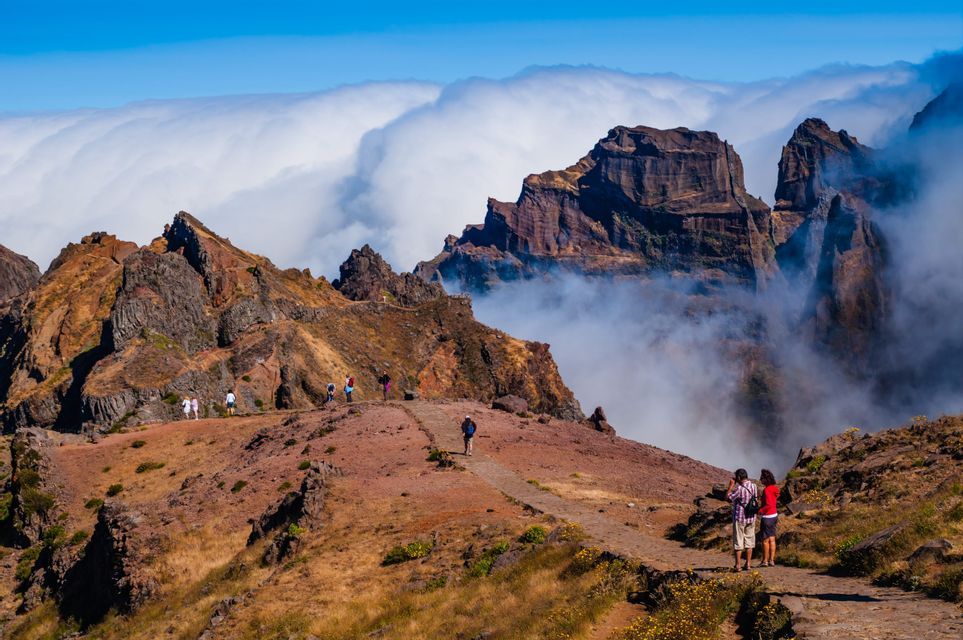 A WeRoad group trip hikes along a winding, rocky mountain path with jagged peaks rising above a sea of clouds.