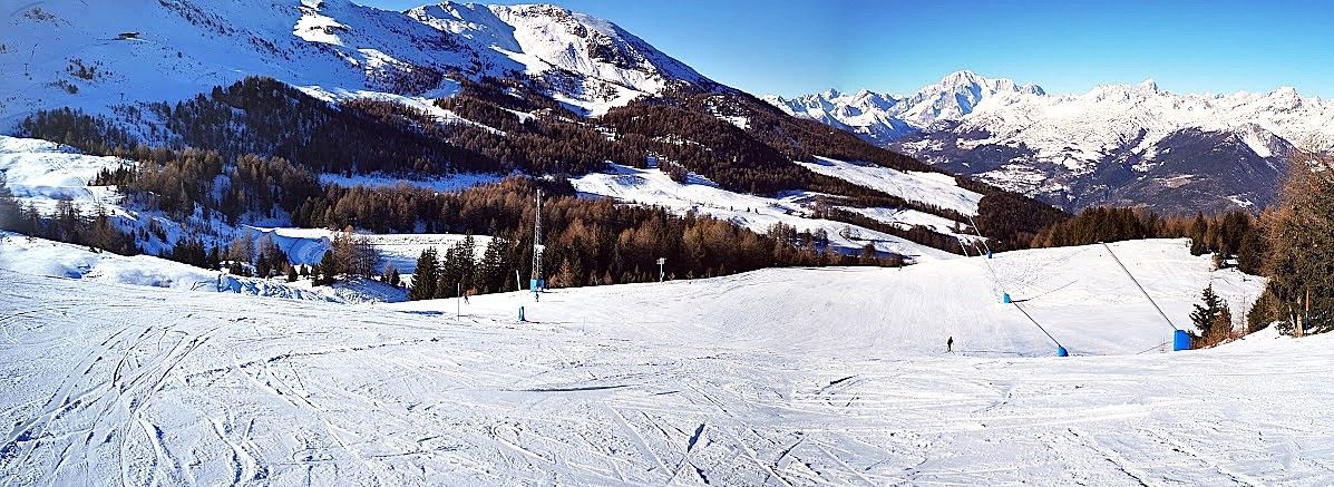 Vista panoramica di un'ampia pista da sci battuta in una giornata di sole, con montagne e foreste innevate sullo sfondo.