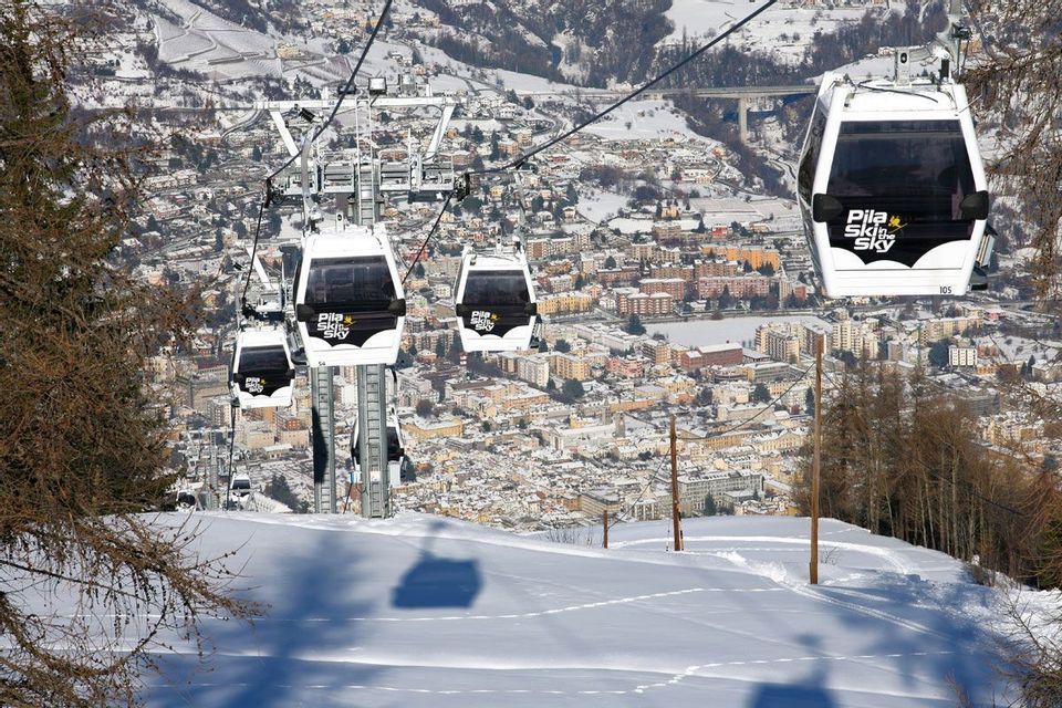 Una fila di bianche funivie sale su un pendio montuoso innevato, affacciandosi su una città nella valle sottostante.