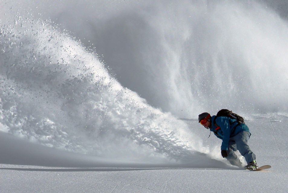 Uno snowboarder con una giacca blu scende intagliando un pendio innevato, sollevando un'ampia nuvola di neve polverosa.