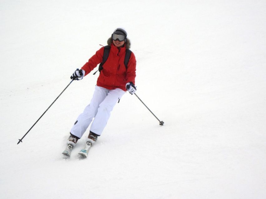 Una persona sorridente con una giacca rossa, pantaloni bianchi e occhiali da sci scia giù per un pendio innevato.