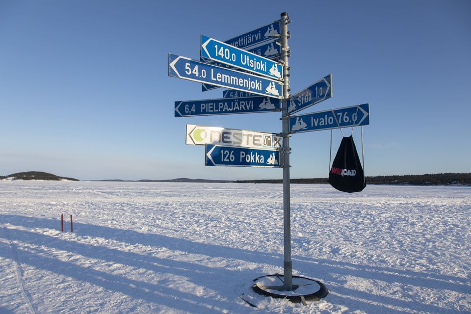 A signpost for snowmobile trails with directions to Ivalo and Utsjoki stands in a vast, snowy landscape under a clear sky.