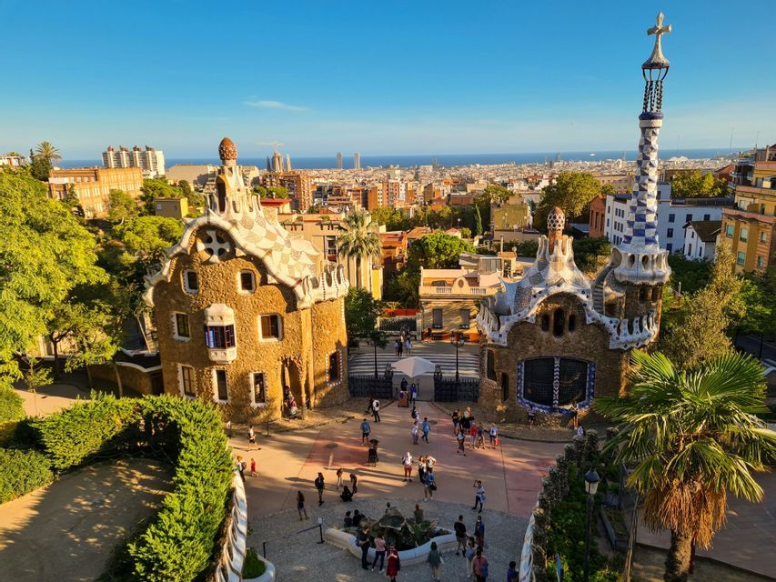 Una vista sopraelevata di due edifici unici, piastrellati a mosaico, con una piazza brulicante di persone, che si affacciano su una città e sul mare.
