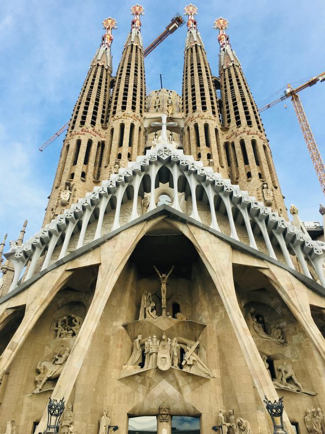 Una ripresa dal basso di una grande cattedrale, con la facciata ornata e le guglie imponenti, in costruzione sotto un cielo blu.