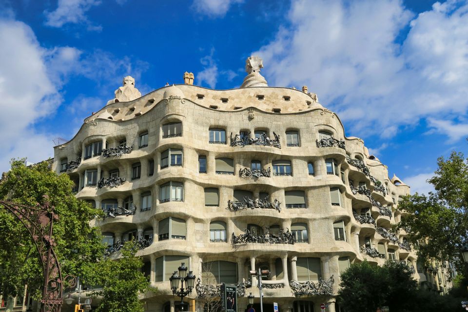 Un grande edificio in pietra con una facciata ondulata e balconi elaborati in metallo sotto un cielo azzurro parzialmente nuvoloso.