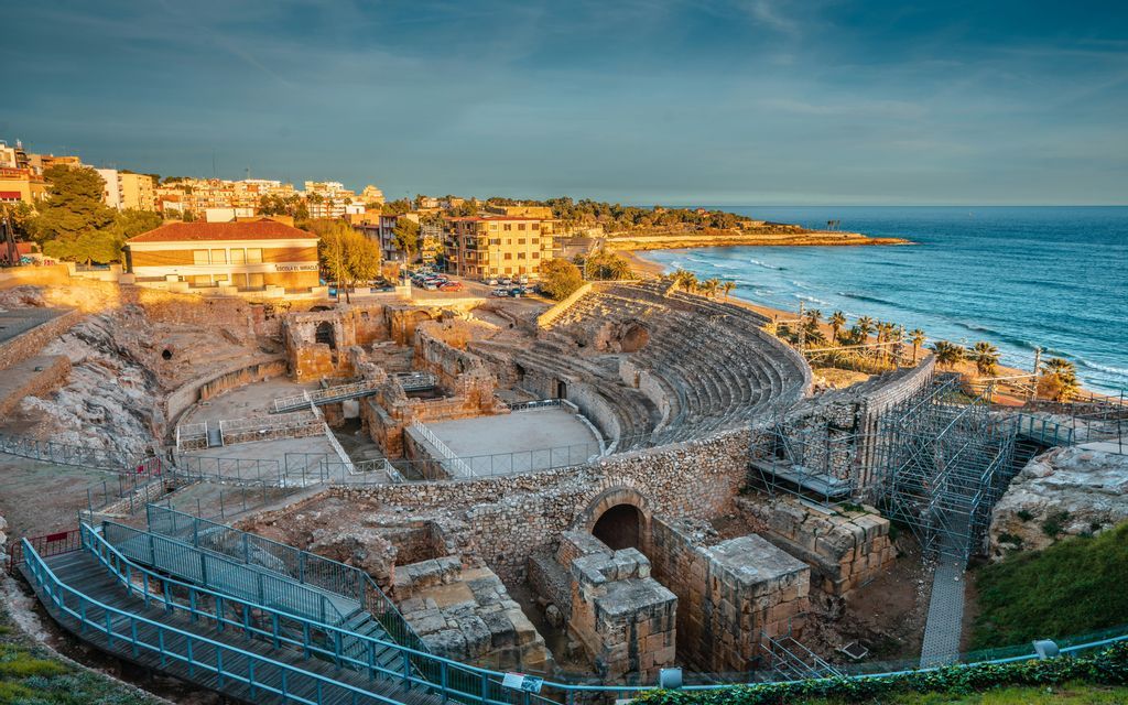 Un antico anfiteatro in pietra si affaccia su una spiaggia sabbiosa e sul mare, con una città moderna sullo sfondo al tramonto.