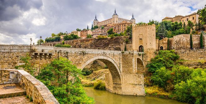 Un ponte ad arco in pietra medievale con una torre fortificata attraversa un fiume ai piedi di una storica città collinare sotto un cielo nuvoloso.