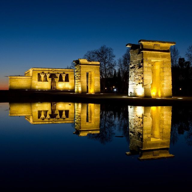 Un antico tempio di pietra in stile egizio e i suoi portali sono illuminati al tramonto, riflettendosi perfettamente in acque immobili.