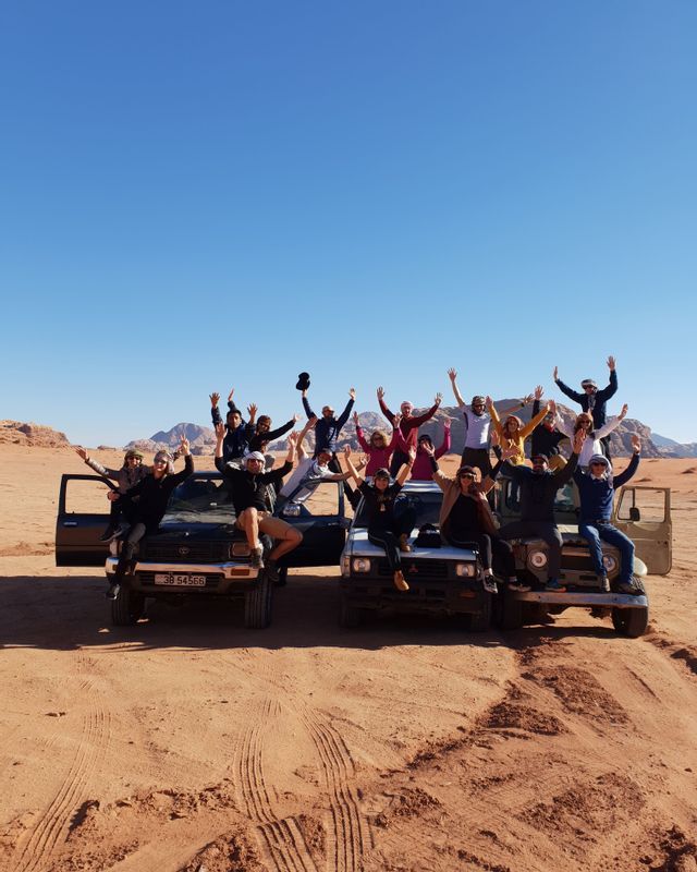 A WeRoad group trip posing with arms raised on three off-road vehicles in a desert landscape under a clear blue sky.