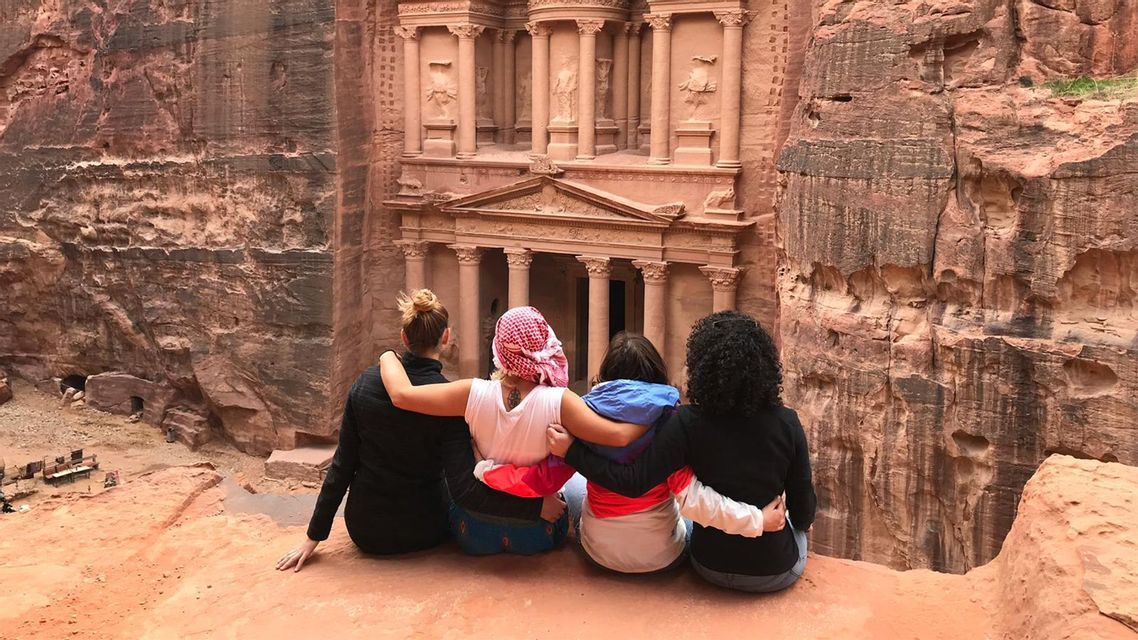 A WeRoad group trip of four women sitting with their arms around each other, looking at an ancient temple carved into a rock cliff.
