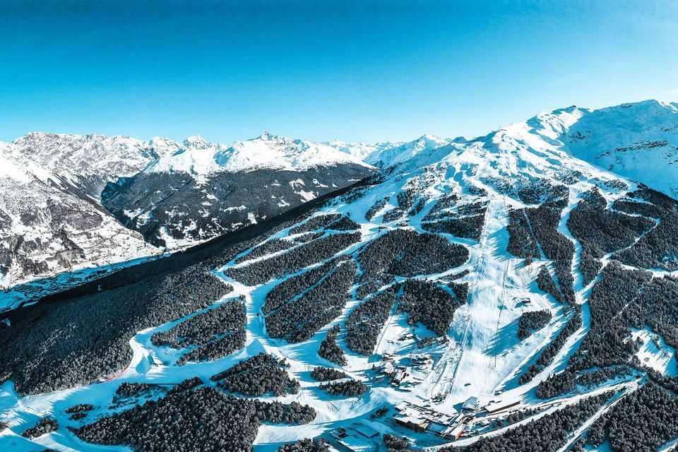 Una vista aerea di una stazione sciistica con diverse piste che attraversano una foresta montana innevata sotto un cielo sereno.