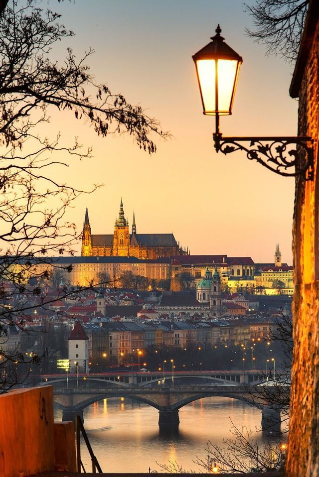 Une ville européenne illuminée avec un château sur une colline et un pont sur une rivière au coucher du soleil, avec un lampadaire allumé au premier plan.