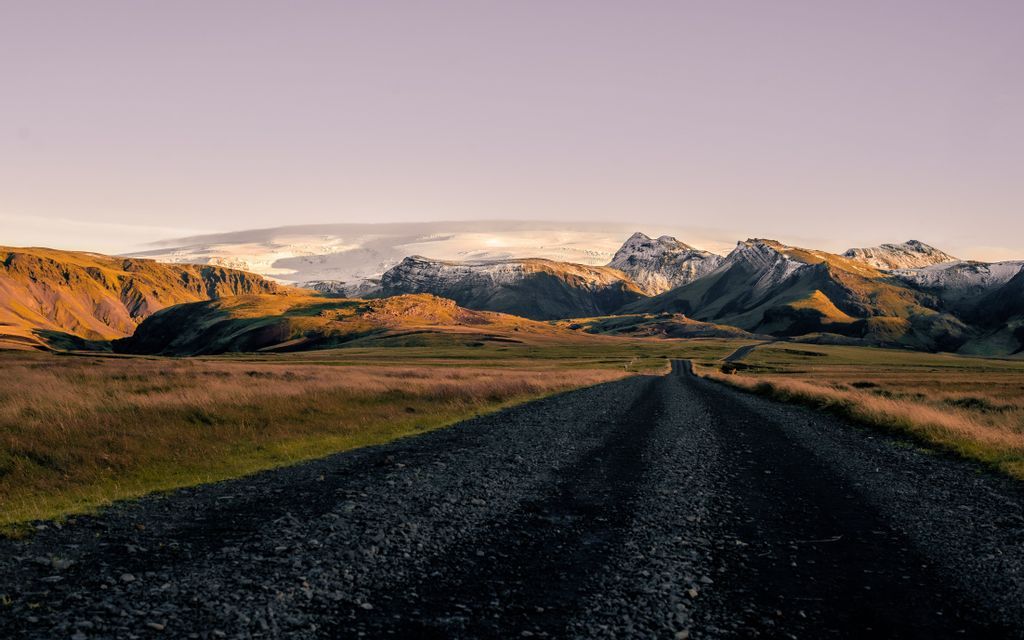 Un camino de grava serpentea por un valle verde hacia montañas soleadas y nevadas y un glaciar lejano bajo un cielo pálido.