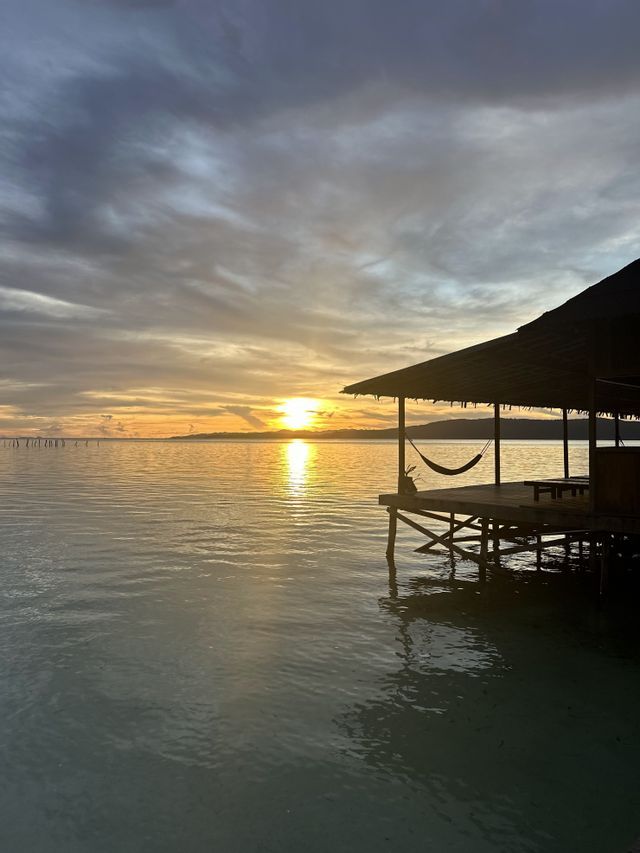 Una hamaca cuelga de una cabaña de madera sobre el agua en calma mientras el sol se pone en el horizonte bajo un cielo nublado.