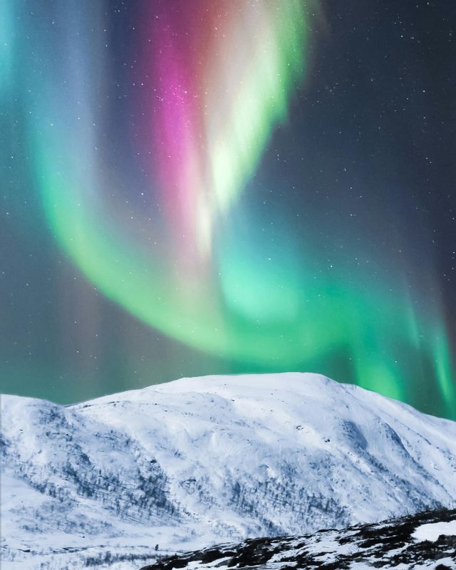 L'aurora boreale con sfumature verdi, rosa e viola si agita nel cielo stellato notturno sopra una montagna innevata.