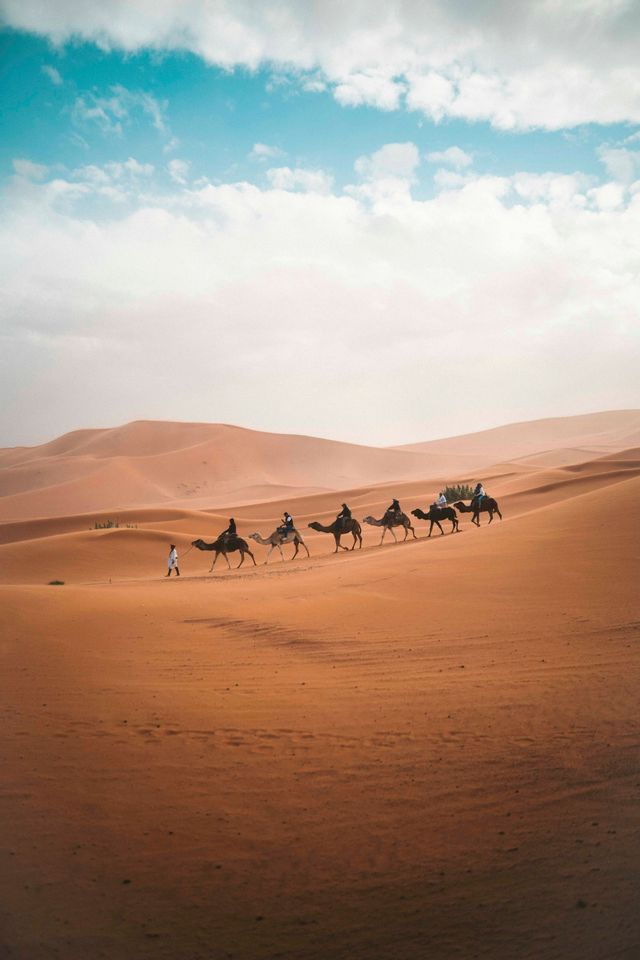 Un viaggio di gruppo WeRoad in cammello, in carovana attraverso vaste dune di sabbia nel deserto sotto un cielo parzialmente nuvoloso.