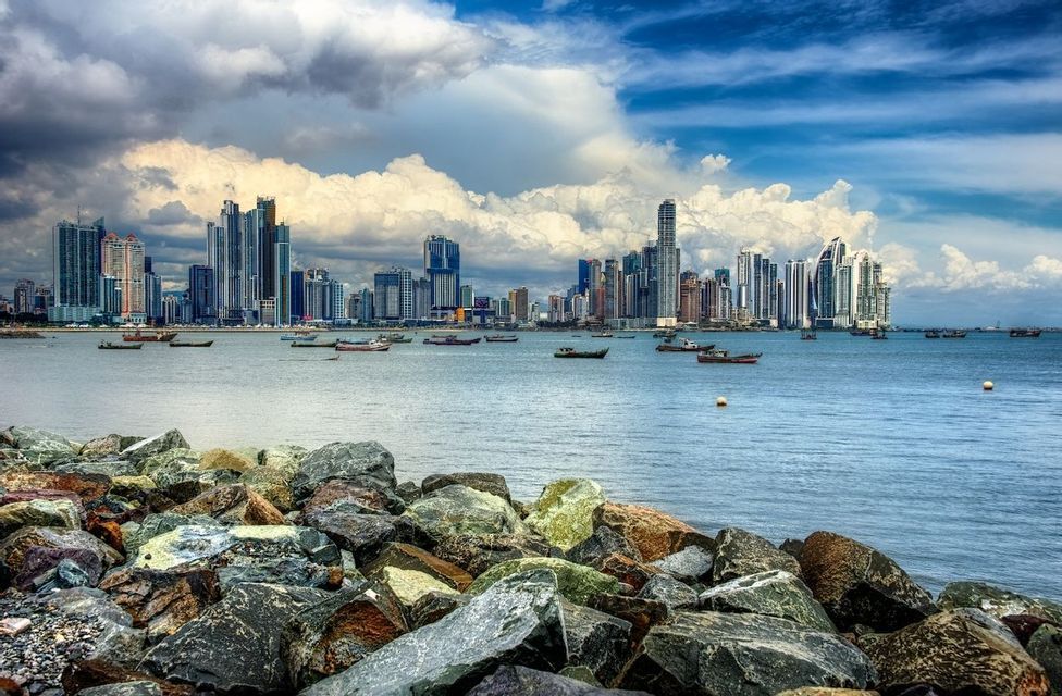 Uno skyline cittadino moderno visto da una costa rocciosa, con piccole imbarcazioni che galleggiano sull'acqua sotto un cielo parzialmente nuvoloso.