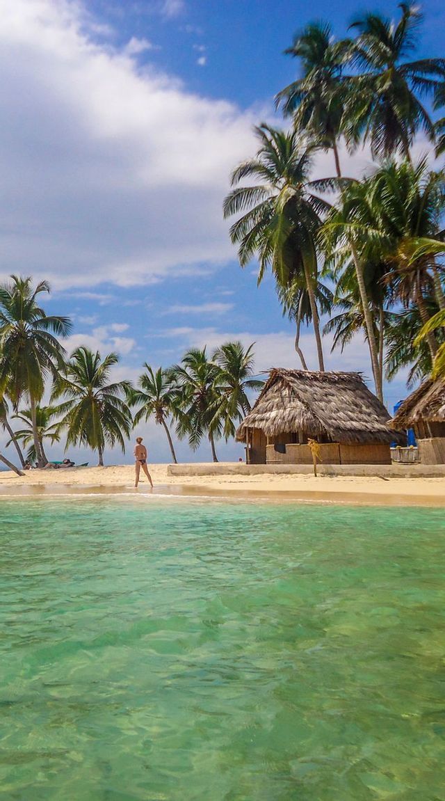 Una persona cammina su una spiaggia di sabbia bianca con acqua turchese cristallina, circondata da palme e una capanna di paglia sotto un cielo blu.