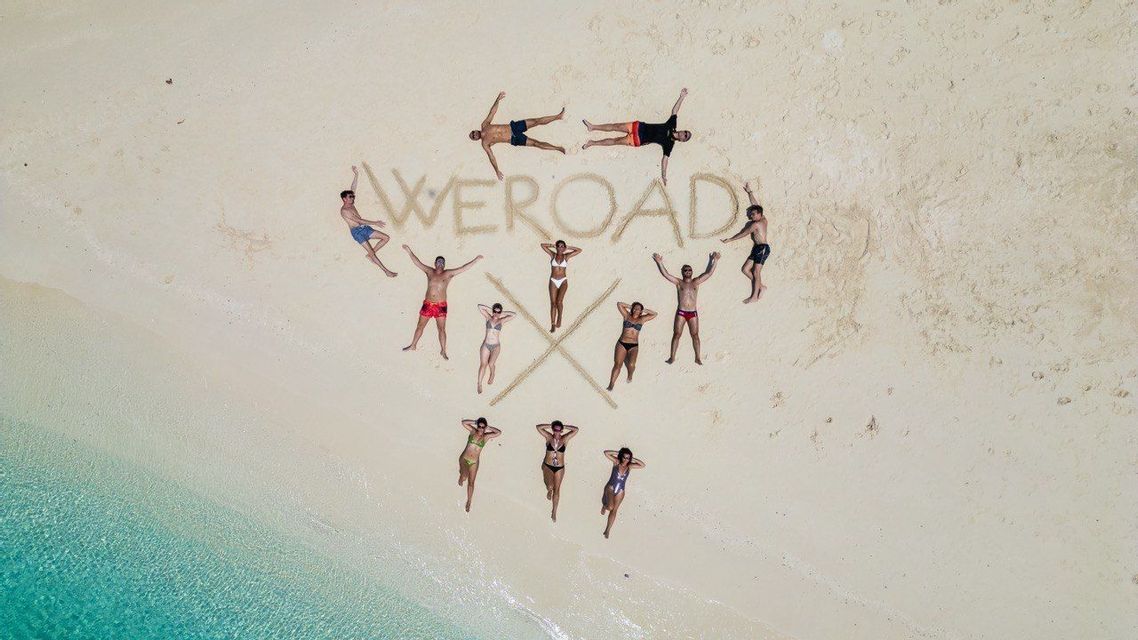 Veduta aerea di un gruppo WeRoad sdraiato su una spiaggia di sabbia bianca attorno alla parola 'WEROAD' scritta sulla sabbia