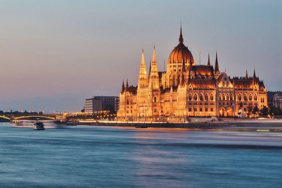 An ornate government building with a dome and spires, brightly illuminated along a river at dusk.