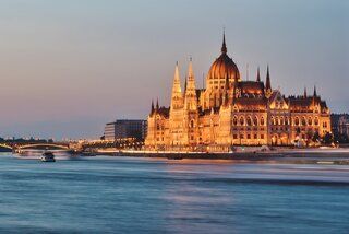 An ornate government building with a large dome and spires, illuminated with golden lights at dusk on a riverbank.