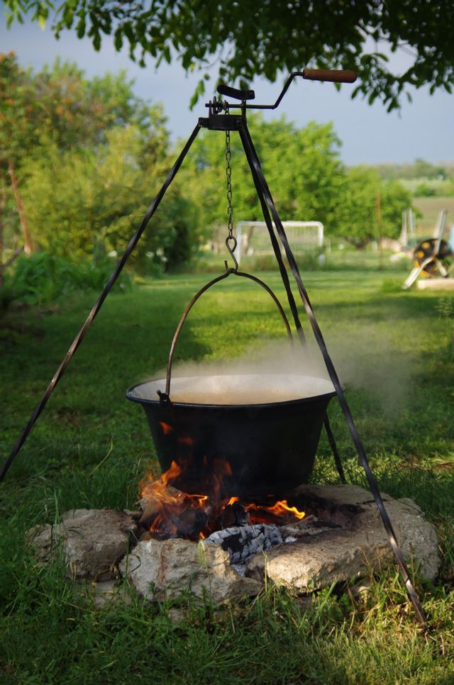 Ein großer schwarzer Kessel dampft über einem offenen Feuer, das in einem Steinkreis auf einer grünen Wiese mit Bäumen im Hintergrund gebaut wurde.