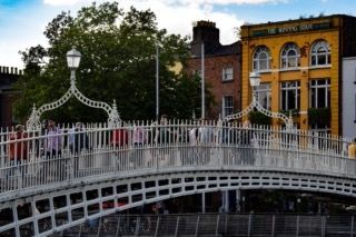 Gente attraversa un affollato e ornato ponte pedonale bianco, con un edificio giallo e alberi verdi sullo sfondo, sotto un cielo azzurro.