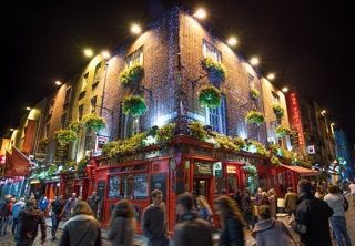 Un pub rosso illuminato in un angolo di un edificio in mattoni di notte, con una folla di persone che cammina per strada.