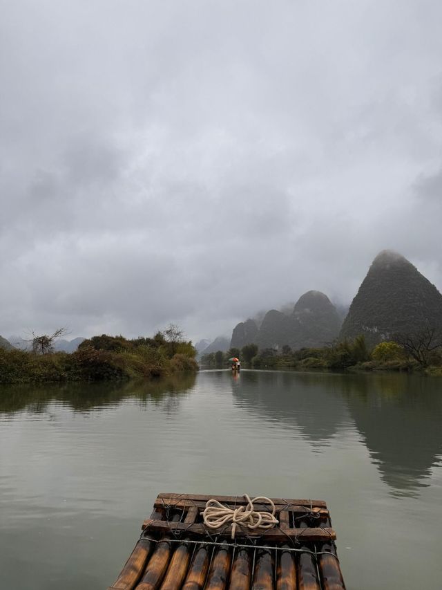 La parte anteriore di una zattera di bambù con corda arrotolata galleggia su un fiume calmo verso montagne nebbiose sotto un cielo coperto.