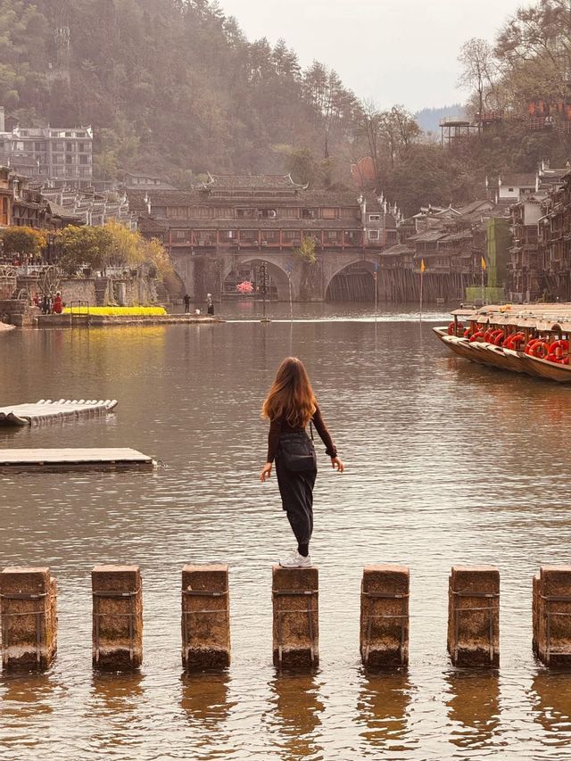 Una donna di spalle alla fotocamera si bilancia su pietre a guado su un fiume, con un villaggio tradizionale e un ponte in lontananza.