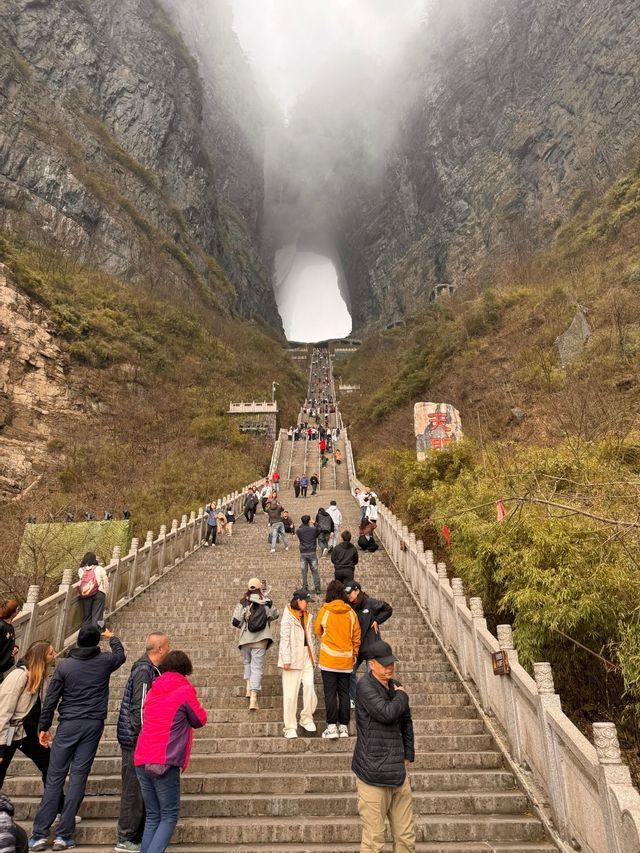 Un gruppo WeRoad in viaggio, con persone che salgono una ripida scalinata in pietra verso un grande arco naturale in una montagna nebbiosa.