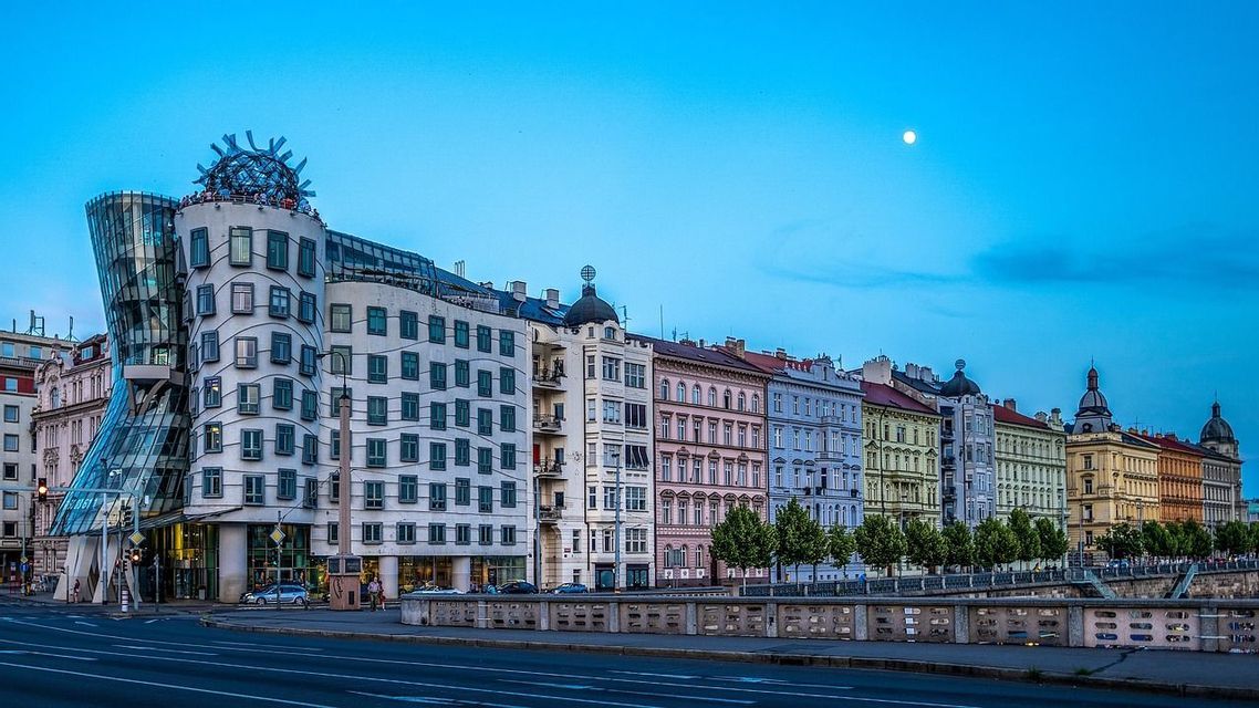 Un edificio unico con architettura in vetro curvo sorge accanto a una fila di edifici tradizionali su una strada cittadina al tramonto.