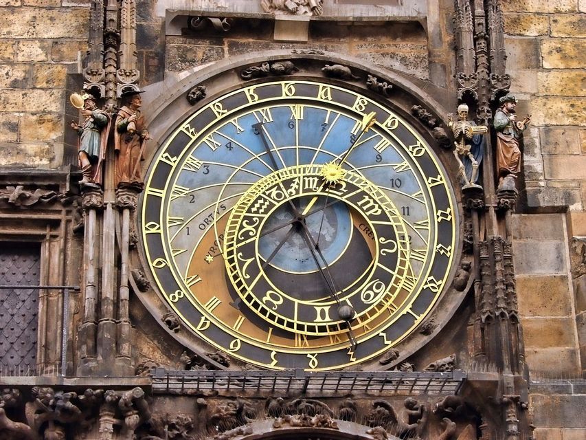 Un orologio astronomico decorato con un complesso quadrante blu e oro, incastonato in un edificio in pietra e affiancato da statue.