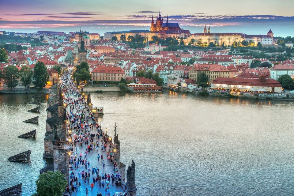 Una vista elevata di un ponte di pietra affollato che attraversa un fiume in una città storica al tramonto, con un castello illuminato su una collina.