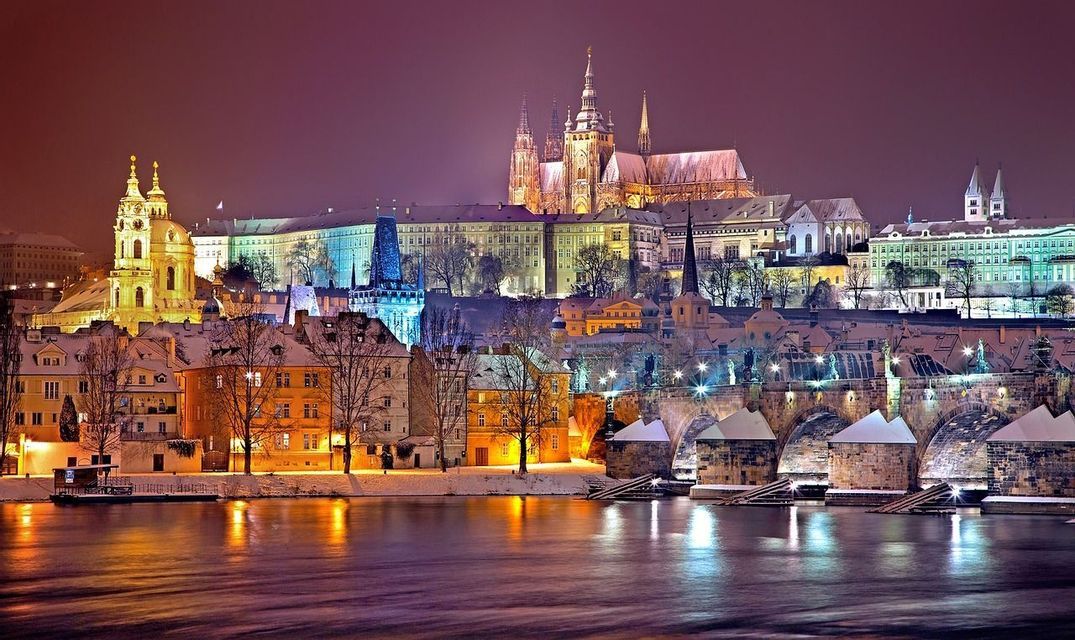 Una città innevata con un grande castello e un ponte di pietra, illuminata di notte con luci che si riflettono nel fiume.