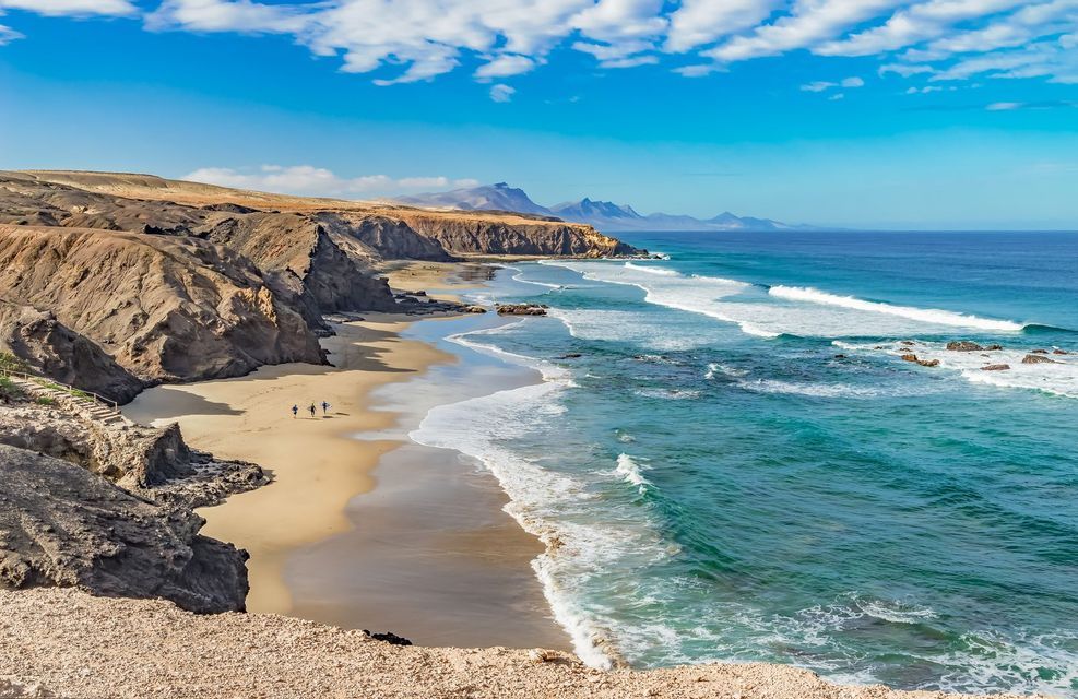 Tre persone di un viaggio di gruppo WeRoad con tavole da surf camminano su una spiaggia sabbiosa accanto a scogliere rocciose e onde turchesi dell'oceano.
