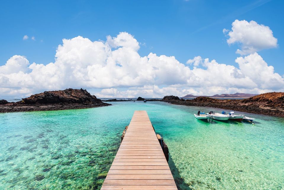 Un pontile di legno si estende su acque calme, limpide e turchesi, con piccole imbarcazioni ormeggiate vicino a rive rocciose sotto un cielo parzialmente nuvoloso.