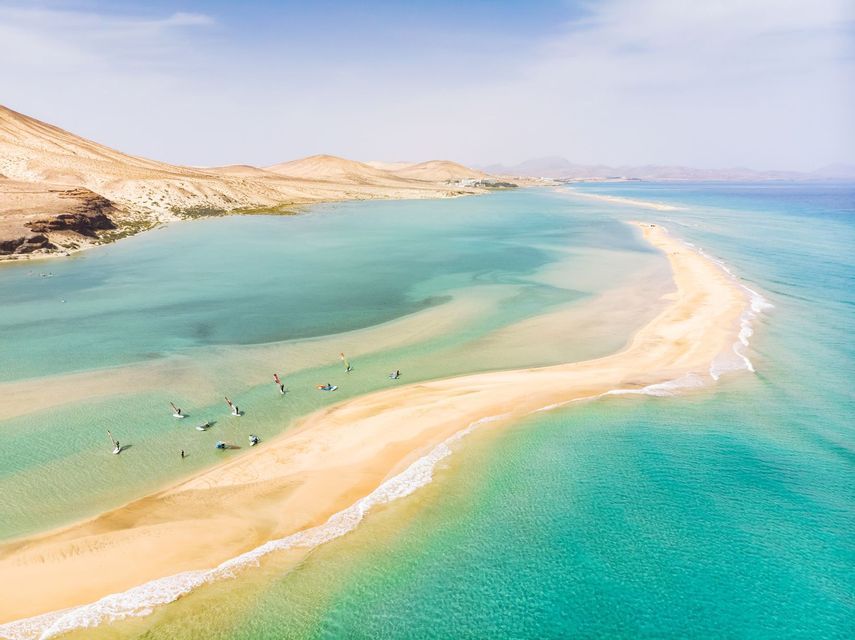 Veduta aerea di un gruppo di persone che fanno windsurf in una laguna turchese vicino a una lunga lingua di sabbia, con colline aride sullo sfondo.