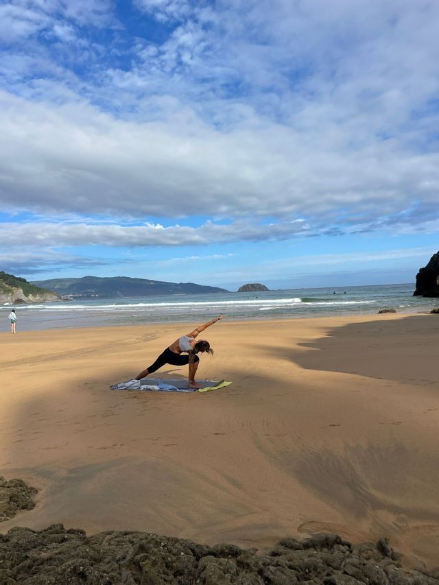 Una donna in posa yoga su un tappetino su una spiaggia sabbiosa, con l'oceano e le colline dietro di lei sotto un cielo parzialmente nuvoloso.