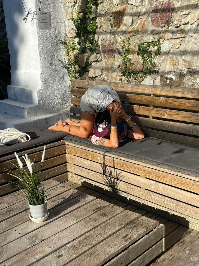 Una donna si inginocchia in una posizione di piegamento in avanti su una panca di legno imbottita su un patio esterno soleggiato.