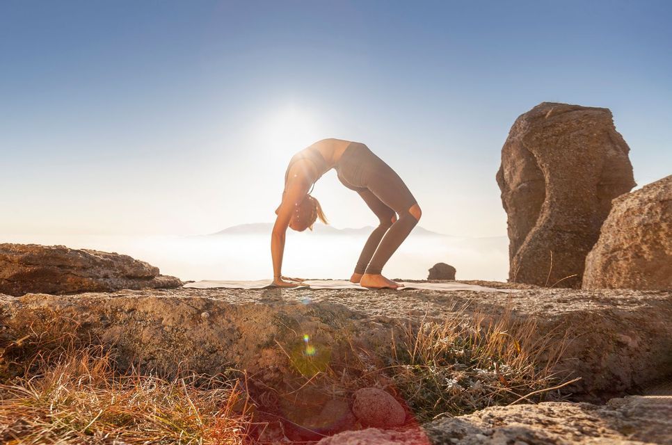 Una donna in posa yoga di inarcamento su un tappetino, su una cima rocciosa, con il sole che splende dietro di lei su un mare di nuvole.