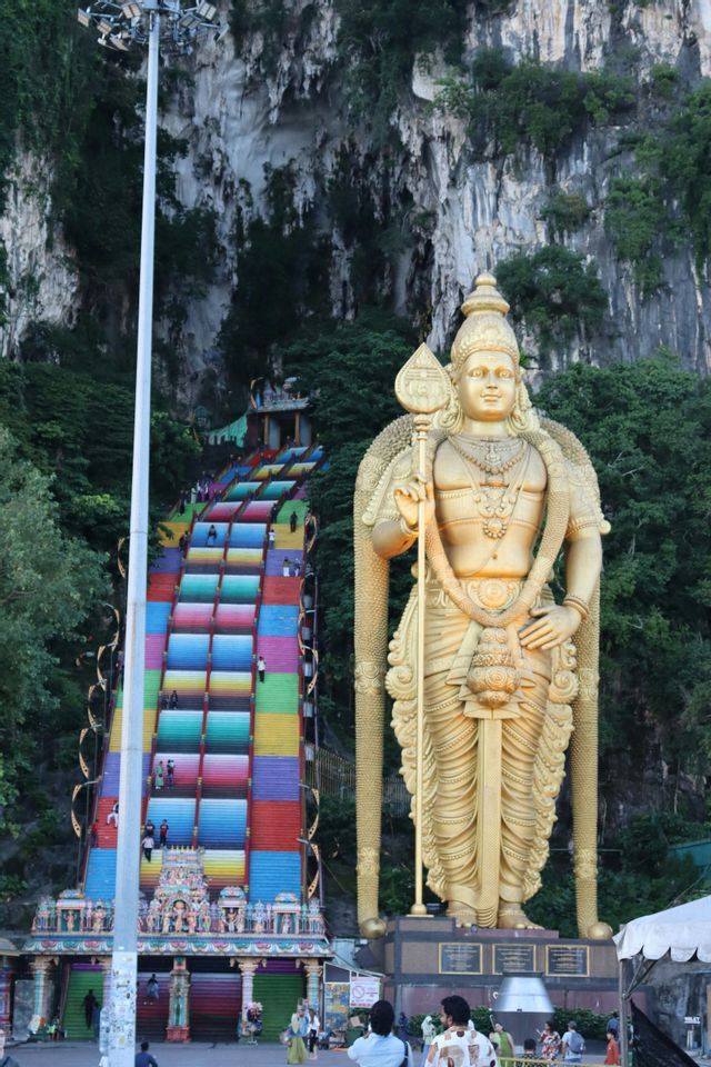 Une statue dorée géante d'une divinité se dresse le long d'un long escalier coloré qui monte une falaise rocheuse, avec des gens marchant sur les marches.