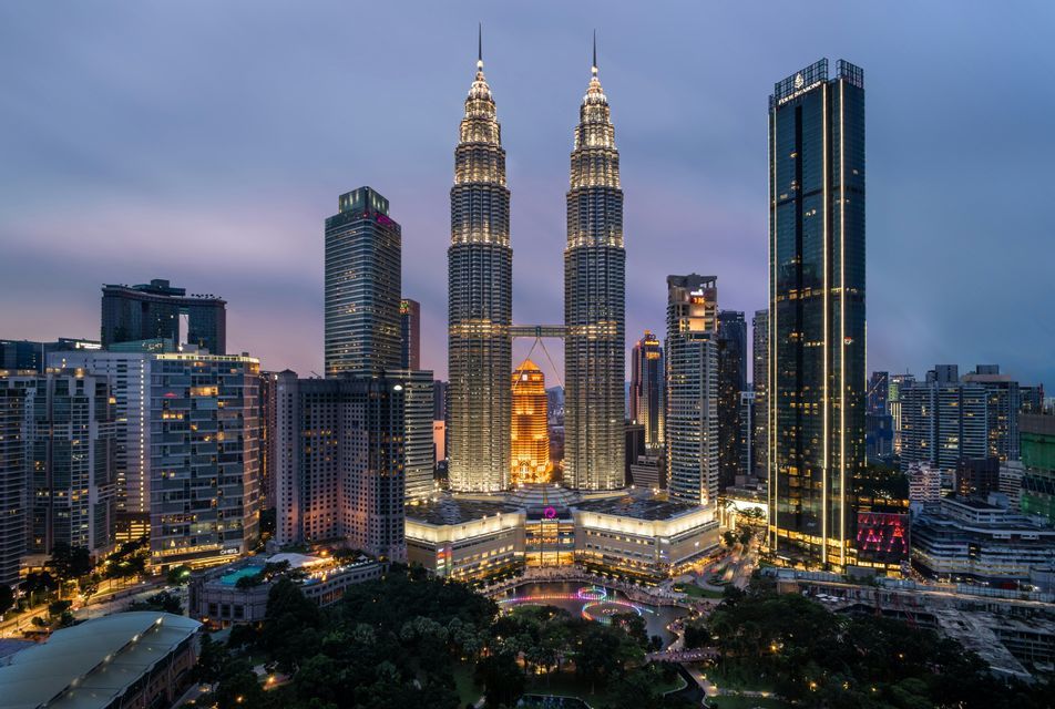 Illuminated twin skyscrapers with a sky bridge stand out in a dense city skyline at dusk.