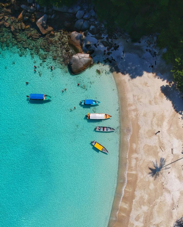 Veduta aerea di barche ormeggiate in limpide acque turchesi, con persone che nuotano vicino a una spiaggia sabbiosa e una costa rocciosa.